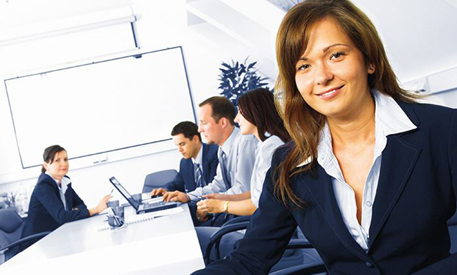 Woman in a business suit smiles at camera while her colleagues meet in the background