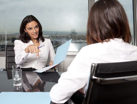  Two business women holding folders sit across from each other at a desk 