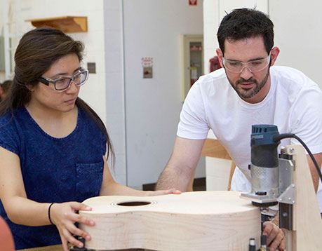 Man and woman sand wooden guitar while wearing safety glasses