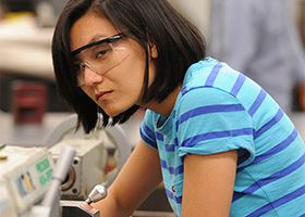 Female student in blue striped shirt and safety goggles leans over manufacturing machine