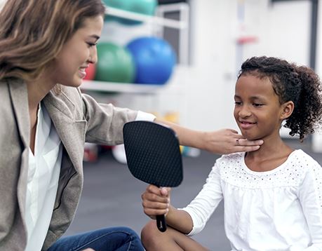 Speech language pathology assistant hold two fingers to a girl's throat as she looks in a mirror